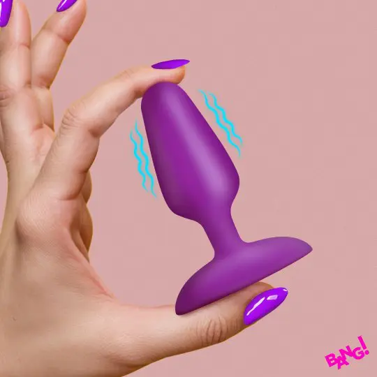 Hand holding a small purple silicone butt plug against a pink background.